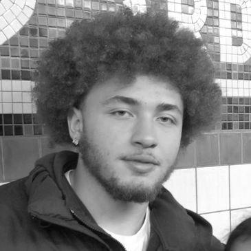 Young man with curly hair and a beard in a tiled indoor setting.