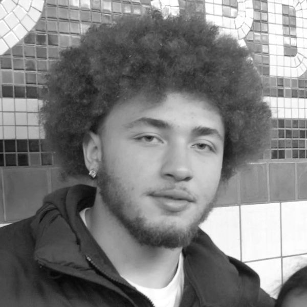 Young man with curly hair and a beard in a tiled indoor setting.