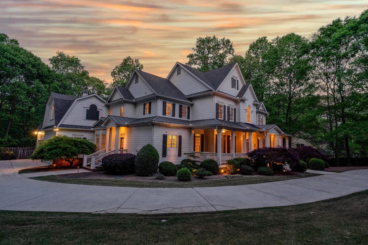 Large white house with warm lights at dusk and surrounding greenery.