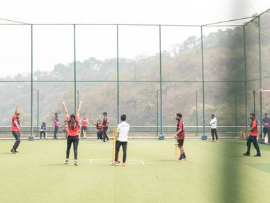 Group playing cricket outdoors on a green field with hills in the background.