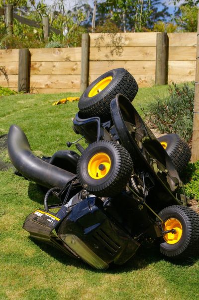 Upside-down lawnmower with yellow wheels on grass near wooden fence.