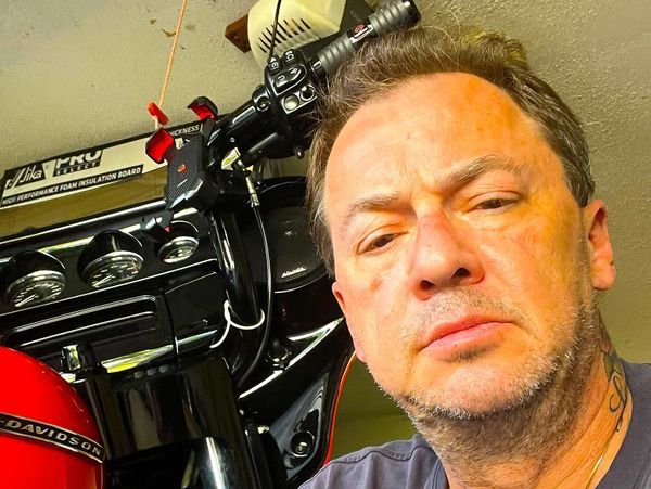 Man with cross necklace taking selfie near a red Harley-Davidson motorcycle.