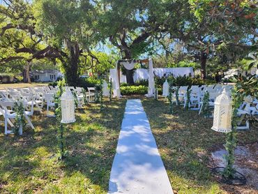 Outdoor wedding setup with white chairs and lanterns along a white aisle.