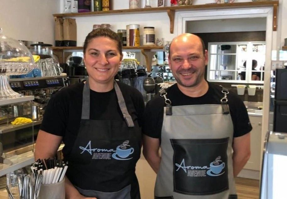 Two smiling baristas wearing Aroma Avenue aprons in a cozy coffee shop.