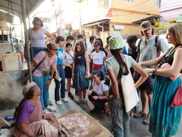 A diverse group of people gathered around an elderly woman selling traditional snacks on the street.