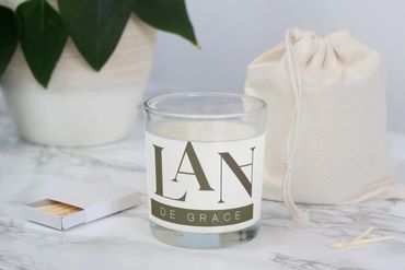 A scented candle labeled 'Lan De Grace' on a marble surface with matches and a plant nearby.