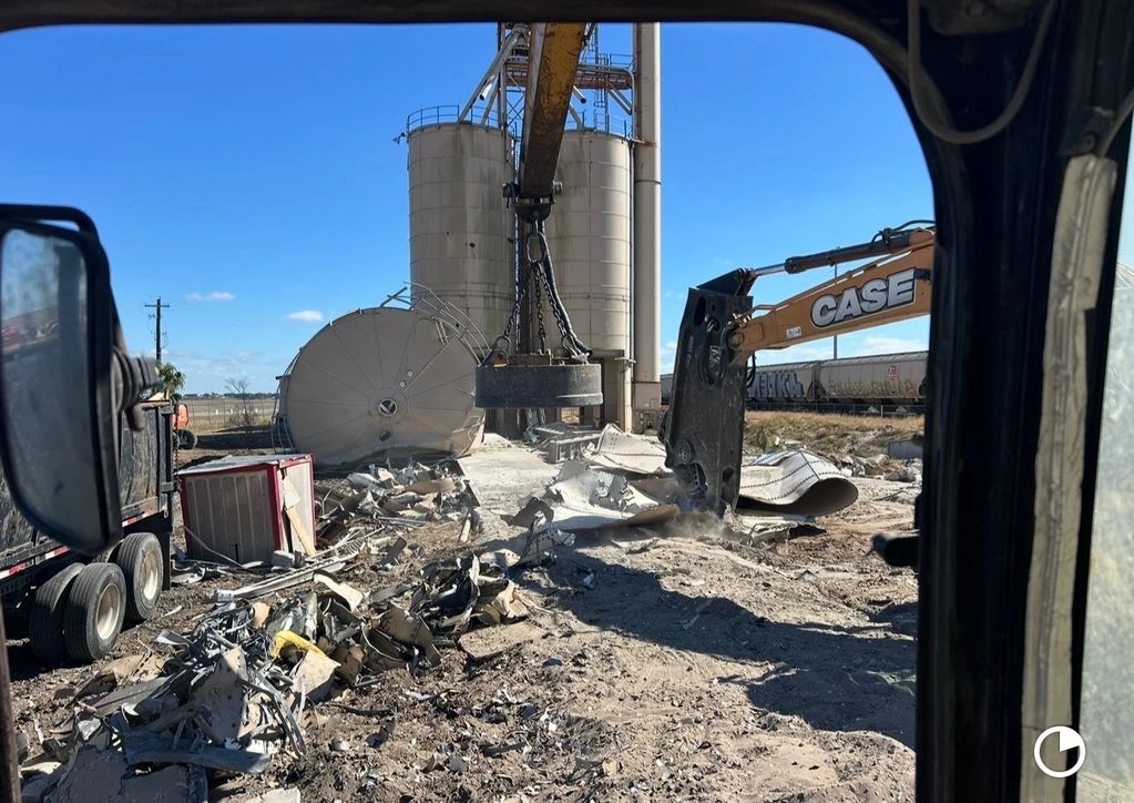 Construction equipment demolishing large silos under a clear blue sky.