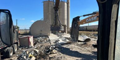 Excavator demolishing large industrial silos in a dusty construction site.