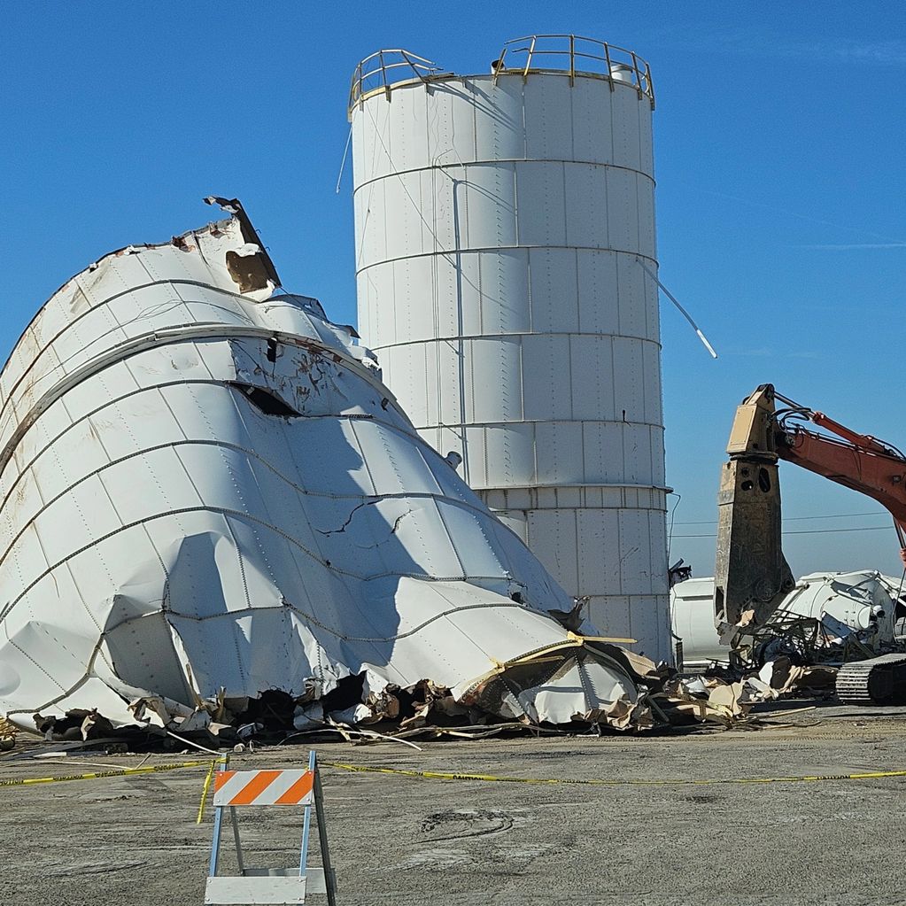 Demolition of large white industrial silos with heavy machinery under a clear blue sky.