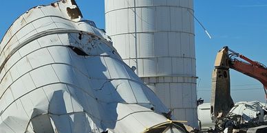 Collapsed metal silo next to intact silo with construction equipment nearby.