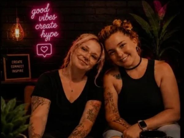 Two smiling women with tattoos sit close together under neon signs.