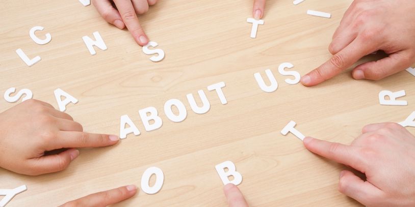 Fingers pointing at letters spelling "ABOUT US" on a wooden table.