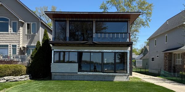 Front elevation of contemporary two-story lakefront renovation in Walled Lake with oversized windows, clean lines, and integrated wood-clad balcony.
