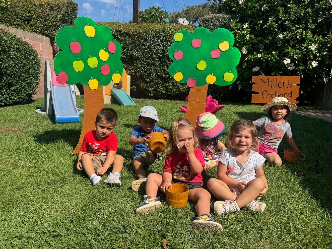 Children sitting on grass in a playful orchard setting with cardboard trees and a sign.
