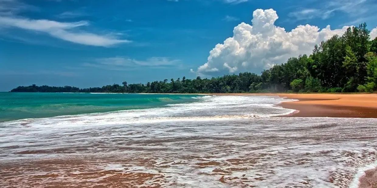A serene beach with turquoise water, sandy shore, and lush greenery under a partly cloudy sky.