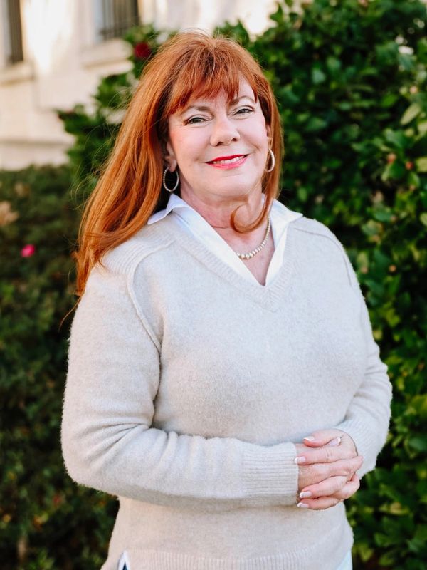 Smiling woman with red hair wearing a beige sweater and blue jeans outdoors.
