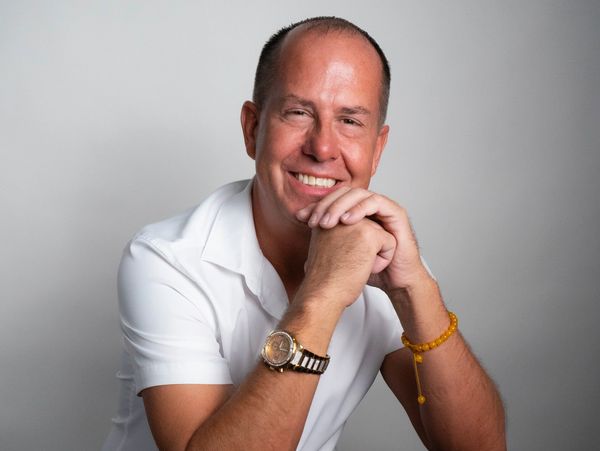 Smiling man in a white shirt and jeans sitting on a stool.