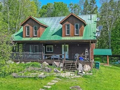 Two-story house with a green metal roof and brown wooden siding, surrounded by greenery, 
