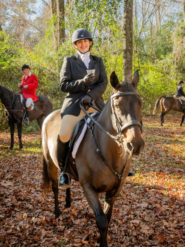 Smiling rider in equestrian attire on a brown horse in a wooded area with autumn leaves.