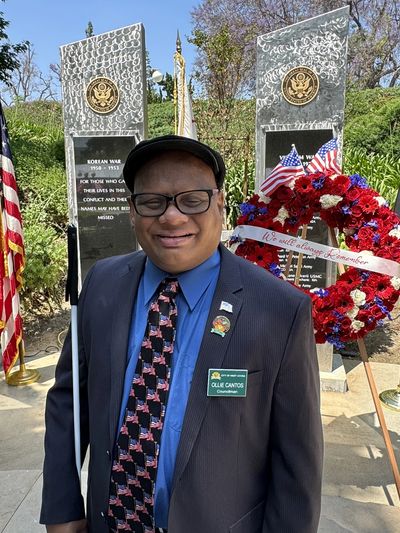 Ollie in a grey suit with a blue shirt and black tie with American flags on it stands before the Vet