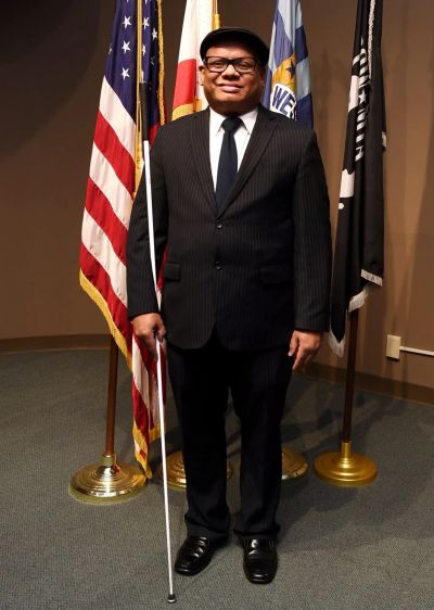 The official photo of City Councilman Ollie Cantos wearing a black suit and holding a white cane.