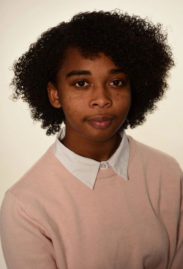 A young woman with curly hair wearing a pink sweater and white collared shirt.