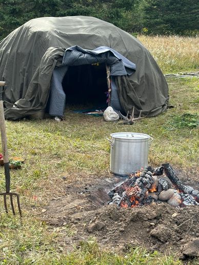 A traditional outdoor sweat lodge with a fire pit and cooking pot in front.