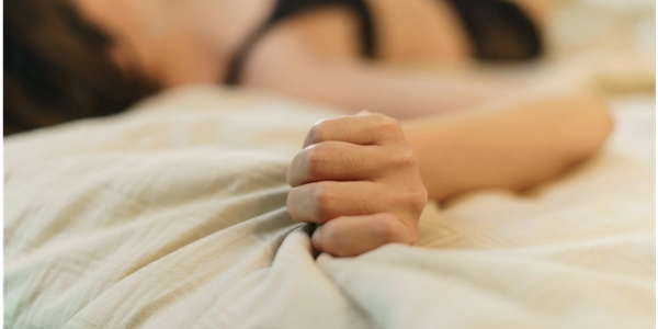 Woman lying on bed, gripping sheets tightly, wearing black lingerie.