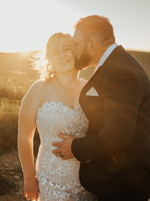 Groom kissing bride on cheek in golden sunset.