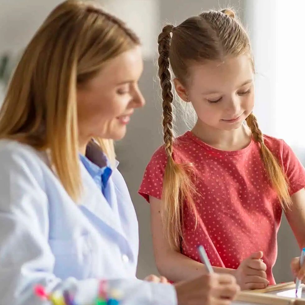 Female teacher helping young girl with homework at a desk.