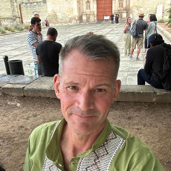 A man in a green shirt takes a selfie outdoors with people and historic buildings in the background.