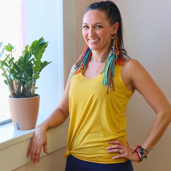 Smiling woman in yellow top with colorful braided hair stands by a window with a plant.
