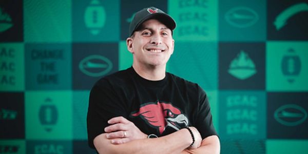Man wearing a black shirt and cap with a red bird logo, smiling with arms crossed.