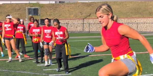 Women playing flag football on a sunny field, one sprinting forward.