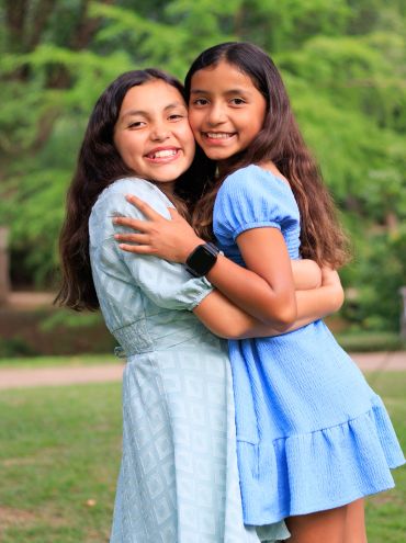 Two girls happily hugging outdoors in pastel dresses.
