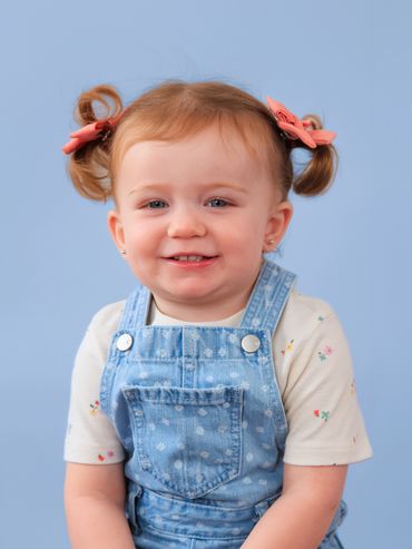 Smiling toddler girl with pigtails and pink bows in denim overalls.