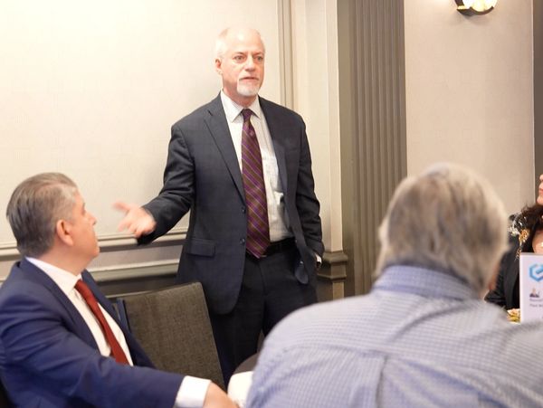 A man in a suit speaks to seated colleagues at a professional meeting.