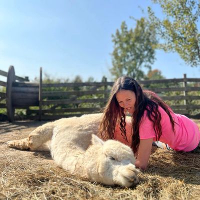 A woman in a pink shirt lies beside a resting alpaca on straw.