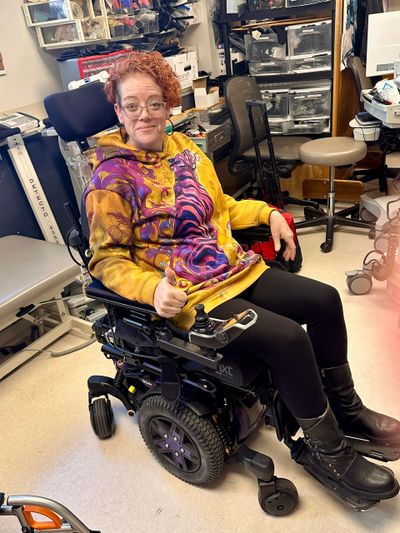 Person smiling and giving thumbs up while sitting in a motorized wheelchair indoors.