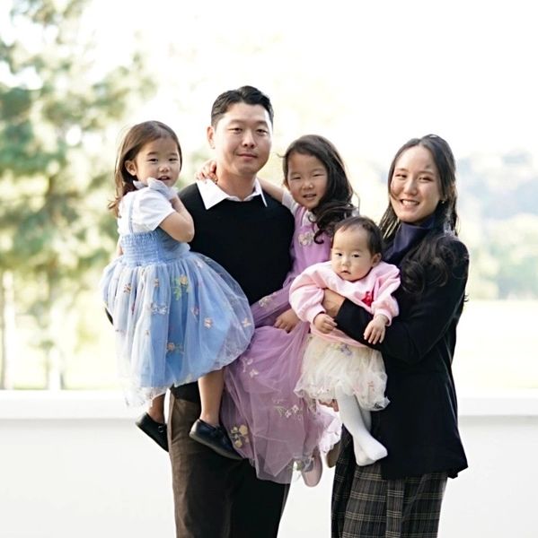 A happy family of five posing outdoors with the parents holding their three young daughters.