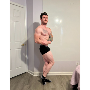 Muscular man with tattoos posing indoors in black shorts and socks.