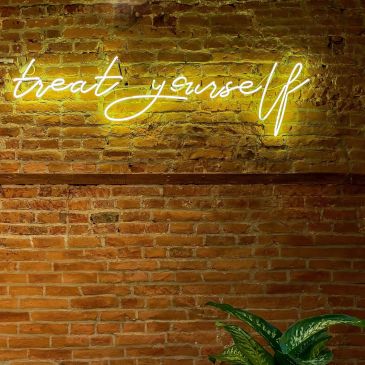 Leuchtendes Schild sagt "treat yourself" an einer Backsteinwand.