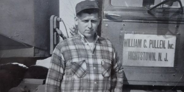 Man in plaid shirt and cap stands by truck with company sign.