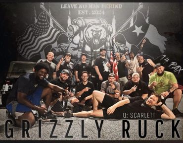 Group of 14 men posing in front of a grizzly bear mural with flags and text.