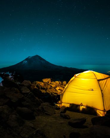 Glowing yellow tent under starry night near a mountain.