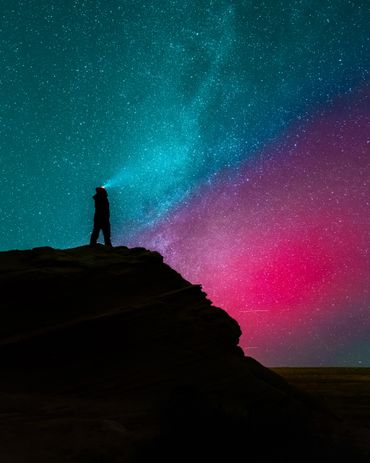 A person with a headlamp stands on a rocky cliff beneath a vibrant starry sky.