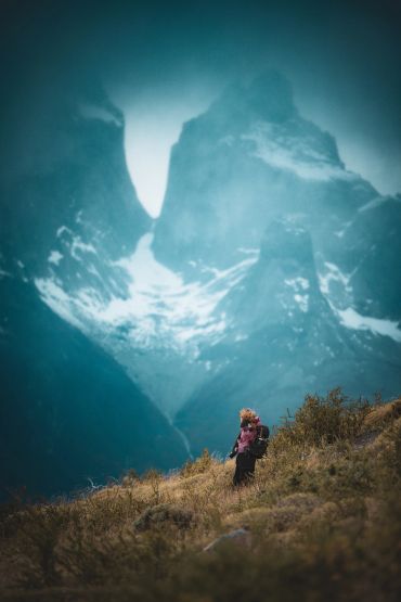A lone hiker stands in a grassy field with towering snowy mountains in the background.