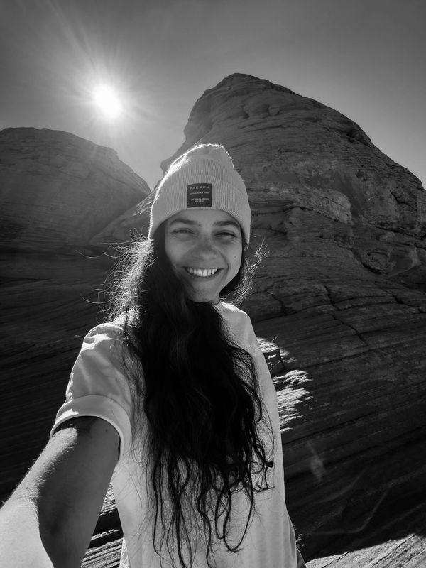 Smiling woman in a beanie takes a selfie against rocky cliffs under bright sun.