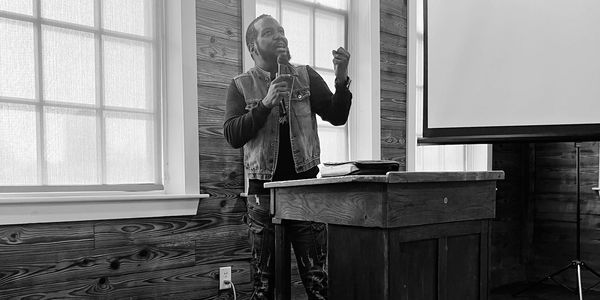 Man speaking with a microphone at a wooden podium in a rustic room.