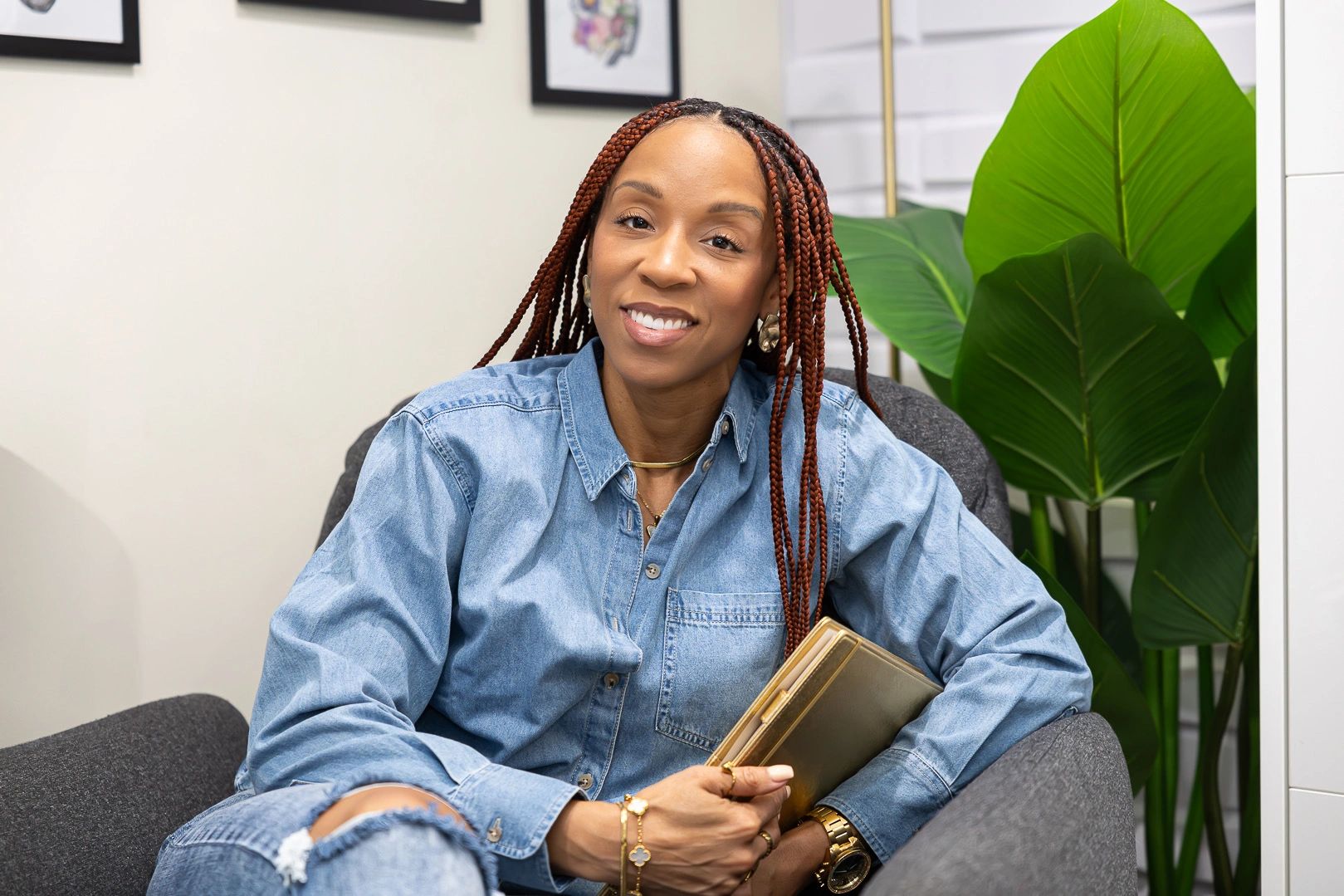 Smiling woman with braided hair wearing a denim shirt and holding a clutch.